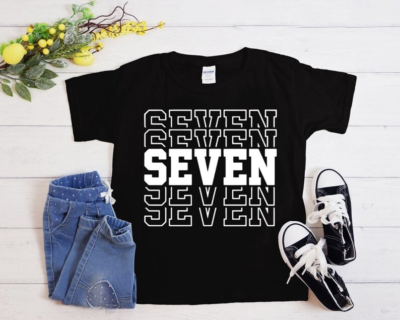 May include: Black t-shirt with white text that says "SEVEN" repeated in a stacked pattern. The text is outlined in black.