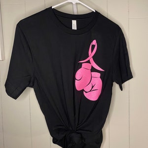 May include: Black t-shirt with a pink ribbon and boxing gloves graphic. The ribbon and gloves are a symbol of support for breast cancer awareness.