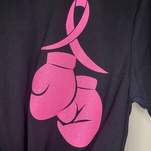 May include: A black t-shirt with a pink ribbon and two pink boxing gloves. The ribbon and gloves are made of glitter.