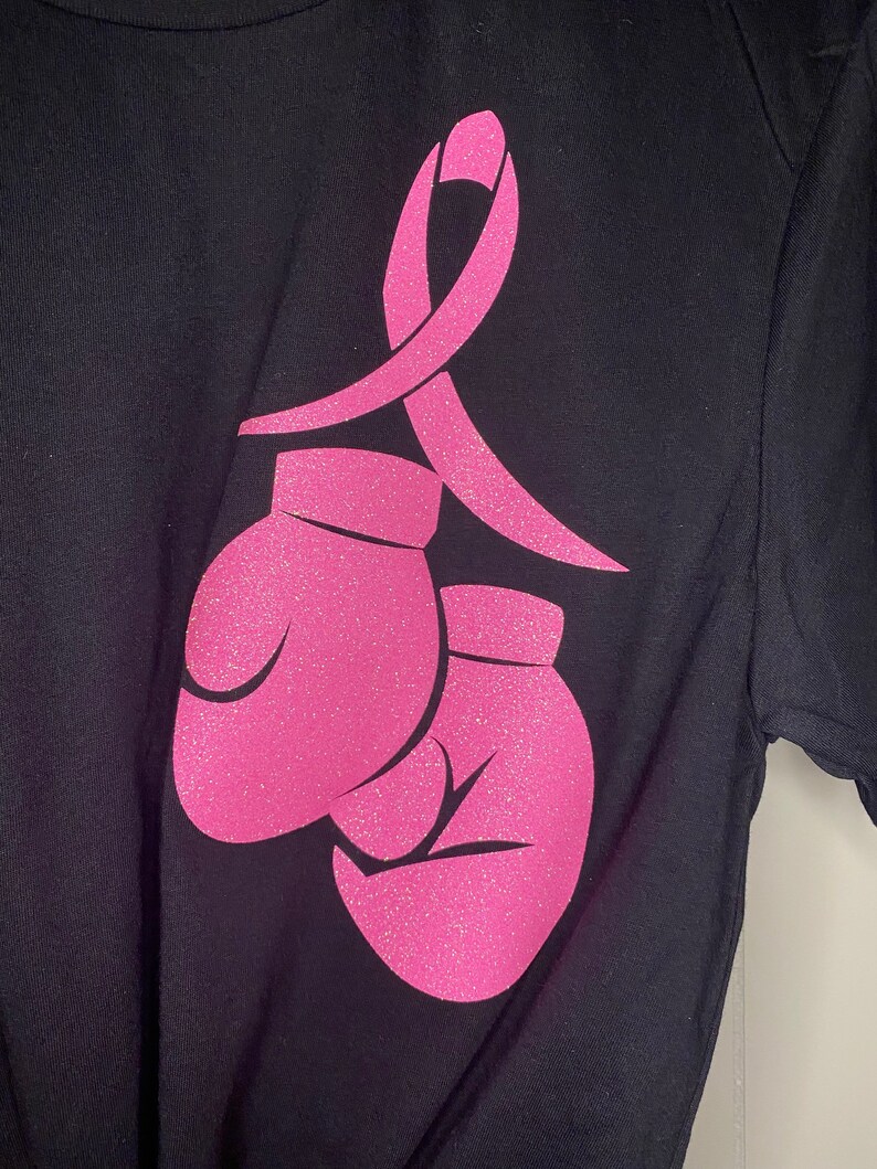 May include: A black t-shirt with a pink ribbon and two pink boxing gloves. The ribbon and gloves are made of glitter.