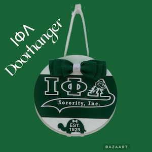 Op de afbeelding: Een ronde deurhanger met een groen en wit ontwerp. De deurhanger heeft de tekst "IΦA Doorhanger" en "Sorority, Inc." in het wit. Er is een groene strik aan de bovenkant bevestigd. Een schildpadgrafiek staat onderaan met de tekst "EST. 1929".