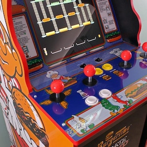 Könnte beinhalten: Ein Vintage-Spielautomat mit farbenfrohem Design. Das Gehäuse verfügt über einen Bildschirm, der ein Spiel anzeigt, rote Joysticks und Knöpfe. Die Seite des Automaten zeigt eine Zeichentrickfigur und die Worte "BurgerTime" unten.