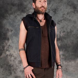 May include: A man wearing a dark blue sleeveless vest with a brown shirt underneath. He is wearing brown cargo pants and has a necklace and bracelets on his wrists.