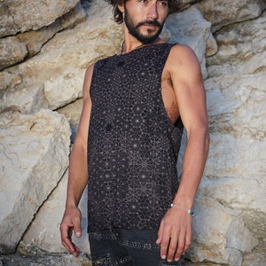 Puede incluir: Un hombre con una camiseta negra sin mangas con un patrón geométrico y pantalones cortos negros. Está de pie frente a un fondo rocoso.