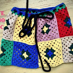 Op de afbeelding: Een kleurrijke gehaakte rok met een zwarte strik sluiting. De rok is gemaakt van vierkanten in verschillende kleuren, waaronder rood, groen, blauw, geel en roze.