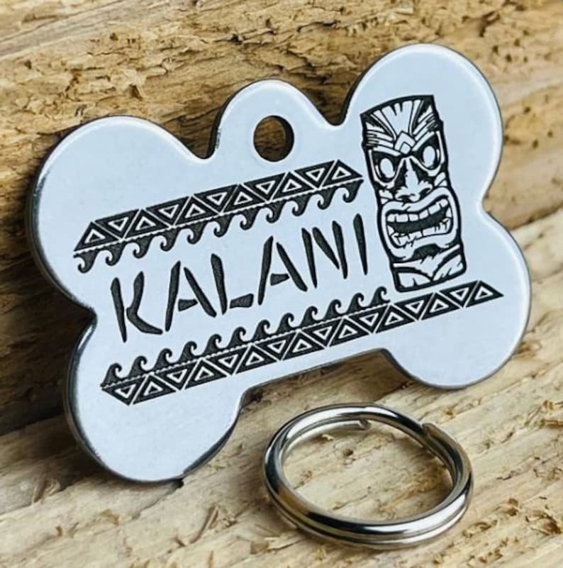 K&ouml;nnte beinhalten: Ein silberner, knochenf&ouml;rmiger Hundeanh&auml;nger mit einem schwarz-wei&szlig;en Design. Der Anh&auml;nger zeigt eine Tiki-Maske und den Namen "KALANI" in einer stilisierten Schrift.
