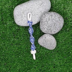 May include: A blue and white braided rope with a white knot at the end. The rope is lying on top of three gray stones, which are on a green artificial grass surface.