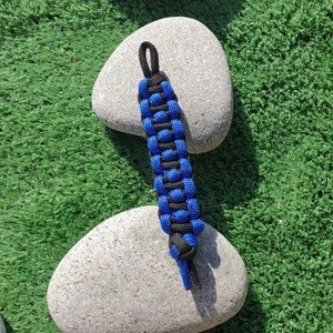 May include: A blue and black paracord bracelet with a knot at the end. The bracelet is sitting on top of two grey stones.