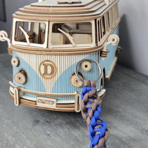 May include: A blue and brown braided keychain with a silver ring. In the background is a wooden model of a blue and white vintage van. The van has the text "SMYIP" on the front.