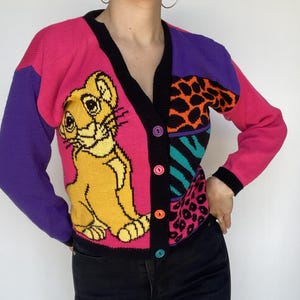 May include: A colorful cardigan sweater featuring a cartoon lion design. The sweater has a V-neck, button closure, and a patchwork design with pink, purple, and black accents. The sleeves are pink and purple. The lion is yellow and the buttons are multi-colored.