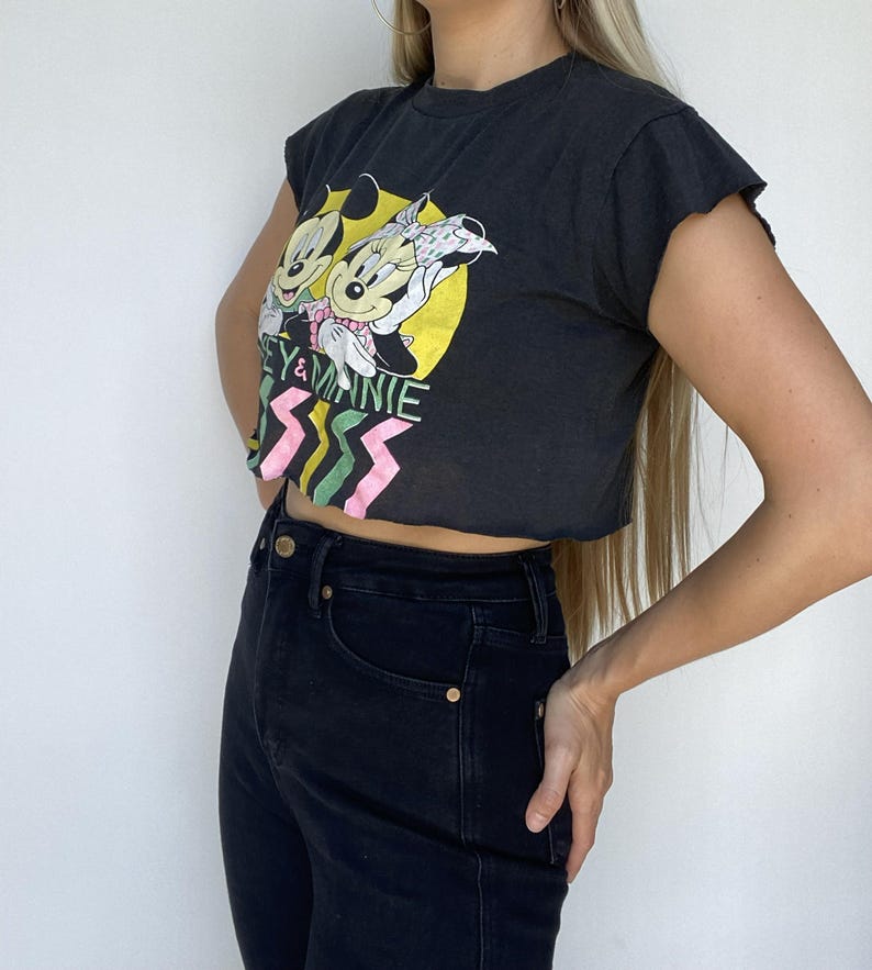 May include: Black crop top featuring Mickey and Minnie Mouse graphic. The graphic includes the text "Mickey & Minnie" with a yellow background and pink and green zig-zag design. The top is paired with black jeans.