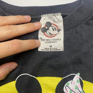 May include: Close-up of a black t-shirt tag with the text "THE WALT DISNEY COMPANY" and a Mickey Mouse graphic. The tag also reads "MADE IN U.S.A." and "FOR CARE SEE REVERSE". The shirt features a Minnie Mouse graphic.