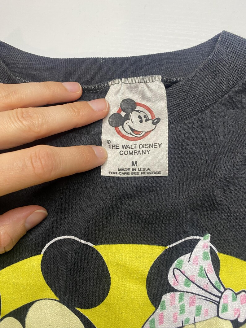 May include: Close-up of a black t-shirt tag with the text "THE WALT DISNEY COMPANY" and a Mickey Mouse graphic. The tag also reads "MADE IN U.S.A." and "FOR CARE SEE REVERSE". The shirt features a Minnie Mouse graphic.