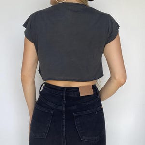 May include: A dark gray, cropped t-shirt with raw edges and short sleeves. The shirt is paired with high-waisted, black denim jeans. The jeans have a classic five-pocket design and a brown leather patch on the back.