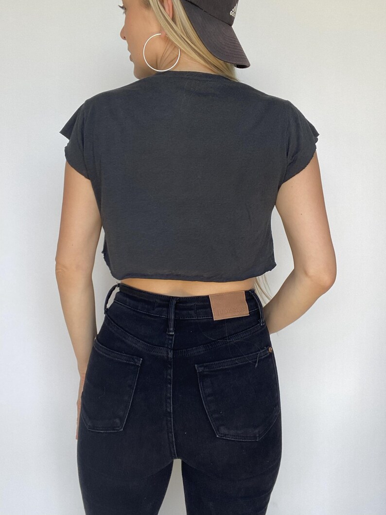 May include: A dark gray, cropped t-shirt with raw edges and short sleeves. The shirt is paired with high-waisted, black denim jeans. The jeans have a classic five-pocket design and a brown leather patch on the back.