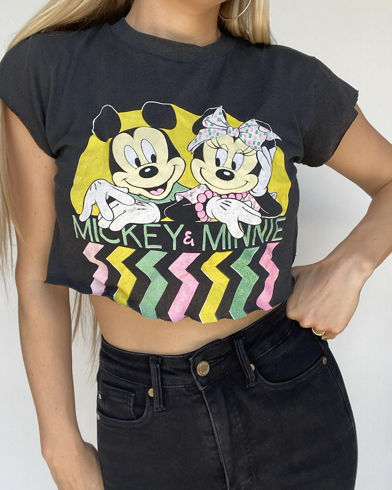 May include: Black crop top featuring Mickey Mouse and Minnie Mouse. The graphic includes the text "Mickey & Minnie" with a colorful zigzag design below the text. The shirt has a distressed look.
