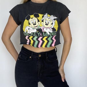May include: Black crop top featuring a graphic of Mickey and Minnie Mouse with the text "Mickey & Minnie." The design includes a yellow background and a colorful zig-zag pattern. The top is paired with black jeans.