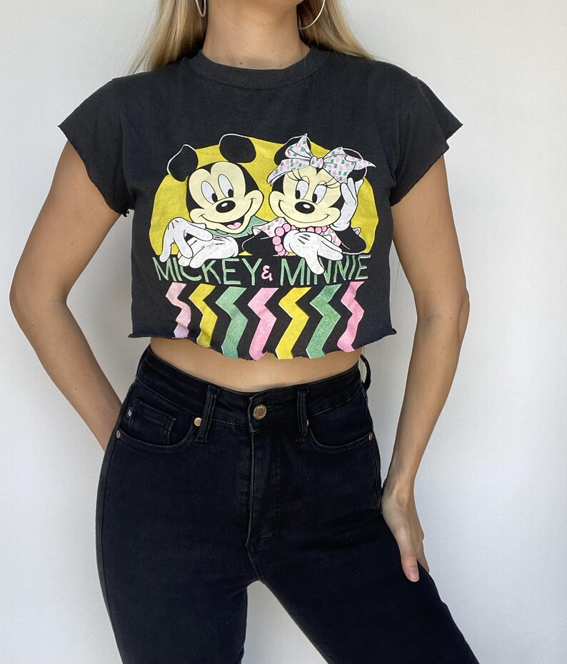 May include: Black crop top featuring a graphic of Mickey and Minnie Mouse with the text "Mickey & Minnie." The design includes a yellow background and a colorful zig-zag pattern. The top is paired with black jeans.