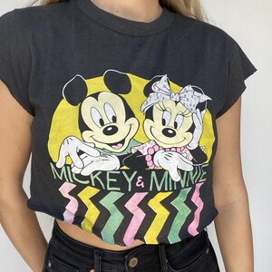 May include: Black t-shirt featuring Mickey and Minnie Mouse. The graphic shows Mickey with a yellow background and Minnie with a pink bow. The text "MICKEY & MINNIE" is above a colorful zigzag pattern.