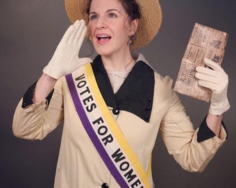 May include: A person wearing a tan coat with black lapels, a straw boater hat, and cream-colored gloves. A sash across the chest features the text VOTES FOR WOMEN in black letters on a purple and yellow background. A printed paper is held in one hand.