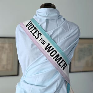 Op de afbeelding: Een lichtblauwe, roze en turquoise sash met de tekst "Votes for Women" in zwart.