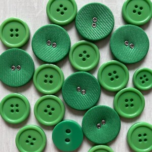 May include: A collection of various green buttons, including round plastic buttons with four holes and fabric-covered buttons with two eyelets. The buttons vary in size and shade of green, creating a textured and visually appealing arrangement.