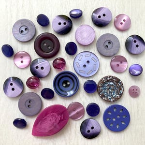 May include: A collection of assorted buttons in various shades of purple, violet, lavender, and deep blue. The buttons feature different shapes, sizes, and textures, including smooth, pearlescent, patterned, and multi-holed designs for sewing.