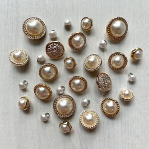 May include: A collection of assorted round buttons in varying sizes, featuring gold-toned metal rims and centres with either smooth cream-coloured faux pearls or textured metallic surfaces. The buttons include both shank and flat-back styles.