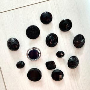 Può includere: Una collezione di vari bottoni neri con diverse trame e design. I bottoni includono superfici sfaccettate, in rilievo e lisce. Un bottone ha un bordo argentato e un centro viola scuro. I bottoni sono disposti su una superficie chiara.