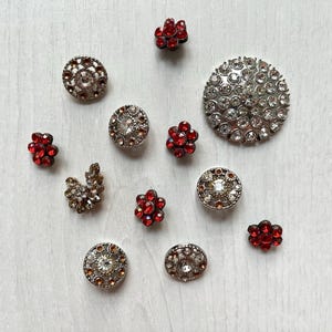 May include: A collection of eleven decorative buttons featuring various designs. Several buttons are circular with clear rhinestones, while others are shaped like small flowers with red rhinestones. One button has a cluster of clear crystals.
