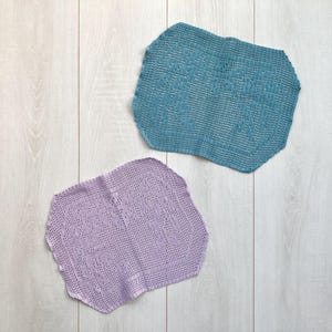 May include: Two handmade, open-weave doilies. One is a light teal colour, and the other is a light purple colour. Both have a decorative, geometric pattern and a scalloped edge. The doilies are placed on a light wood-grain surface.