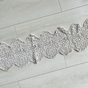 May include: A length of white lace trim with a scalloped edge. The lace features a floral pattern with detailed openwork. The trim is laid diagonally across a light gray wooden surface, showcasing its intricate design.