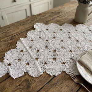 May include: A white, intricately crocheted table runner with a scalloped edge, displayed on a rustic wooden table. The runner's openwork design is complemented by a neutral-toned plate, napkin, and silverware, creating a charming, vintage aesthetic.