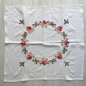 Pode incluir: Toalha de mesa quadrada branca com um design detalhado de ponto cruz. O motivo central é uma coroa de rosas vermelhas, folhas verdes e laços dourados. Acentos florais adicionais de ponto cruz são colocados ao redor da coroa. As bordas são finalizadas com uma delicada renda.