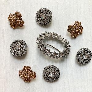 May include: A collection of vintage brooches and pins. The image shows several round, silver-toned pieces with clear crystal accents, gold-toned floral designs, and an oval brooch.