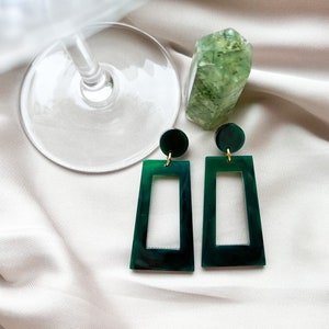 May include: A pair of green rectangular statement earrings with gold accents. The earrings are made of resin and have a marbled effect.