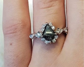 Könnte beinhalten: Ein silberner Ring mit einem schwarzen dreieckigen Diamanten, der von kleineren Diamanten umgeben ist. Der Ring befindet sich an einem Finger.