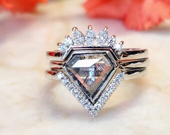 Puede incluir: Un anillo de plata con una gran piedra preciosa de forma de diamante, de color gris, rodeada de diamantes más pequeños. El anillo tiene un diseño delicado en forma de corona con diamantes más pequeños en la banda.