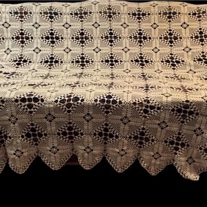 May include: Cream-colored, crocheted sofa cover with a repeating geometric pattern. The cover is draped over a sofa, showcasing its intricate openwork design and scalloped edge. The overall aesthetic is vintage and handmade.