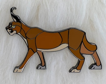 Könnte beinhalten: Eine braune und beigefarbene Emaille-Anstecknadel mit einem stilisierten, gehenden Luchs mit schwarzen Ohren und gelben Augen.