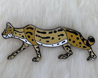 Puede incluir: Un pin de metal dorado de un gato estilizado, caminando, manchado. El gato es marrón, negro y blanco con el vientre blanco.