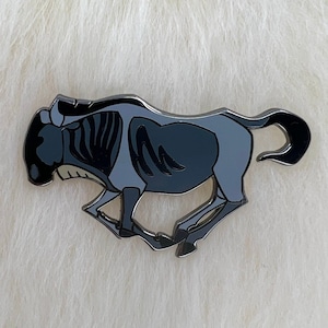 Może przedstawiać: Czarno-szara emaliowana przypinka przedstawiająca biegnącego gnu z kreskówki. Przypinka jest w stylu kreskówkowym i ma błyszczące wykończenie.