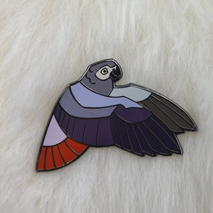 May include: A gray and purple enamel pin of a parrot in flight with red accents on the tail feathers.