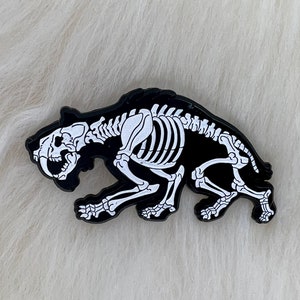 Może przedstawiać: Czarna szpilka emaliowana z białym obrysem szkieletu szablozębnego tygrysa. Kot jest w biegu, z otwartą paszczą i widocznymi kłami.