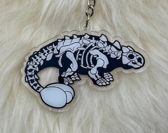 Könnte beinhalten: Ein schwarzer und weißer Skelett-Dinosaurier-Schlüsselanhänger mit einem weißen Ei, das an seinem Schwanz befestigt ist. Der Schlüsselanhänger ist aus klarem Acryl und hat eine silberne Metallkette.