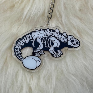 Puede incluir: Un llavero de dinosaurio esqueleto en blanco y negro con un huevo blanco unido a su cola. El llavero está hecho de acrílico transparente y tiene una cadena de metal plateada.