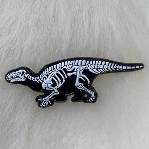 May include: A black dinosaur skeleton pin with white bone details. The pin features a stylized depiction of a dinosaur skeleton, with the bones rendered in white against a black background. The pin is likely made of enamel or a similar material.