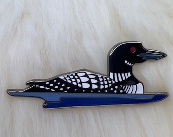 May include: A black and white enamel pin of a loon swimming on a blue lake. The loon has a red eye and a black beak.