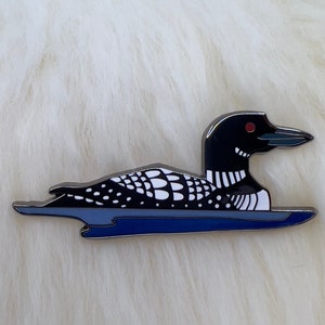 Puede incluir: Un pin de esmalte negro y blanco de un loón nadando en un lago azul. El loón tiene un ojo rojo y un pico negro.