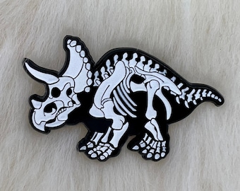 May include: Black and white enamel pin of a dinosaur skeleton, a triceratops, with white details on a black background.